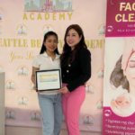 A graduate receives her framed Washington State license from her instructor at Seattle Beauty Academy, standing together in front of the school's branded backdrop