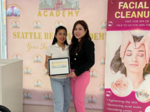 A graduate receives her framed Washington State license from her instructor at Seattle Beauty Academy, standing together in front of the school's branded backdrop