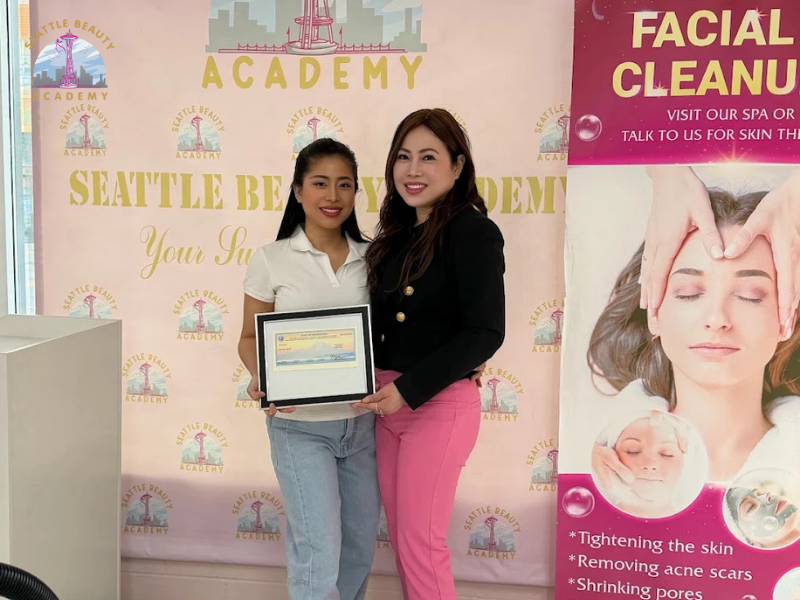 A graduate receives her framed Washington State license from her instructor at Seattle Beauty Academy, standing together in front of the school's branded backdrop
